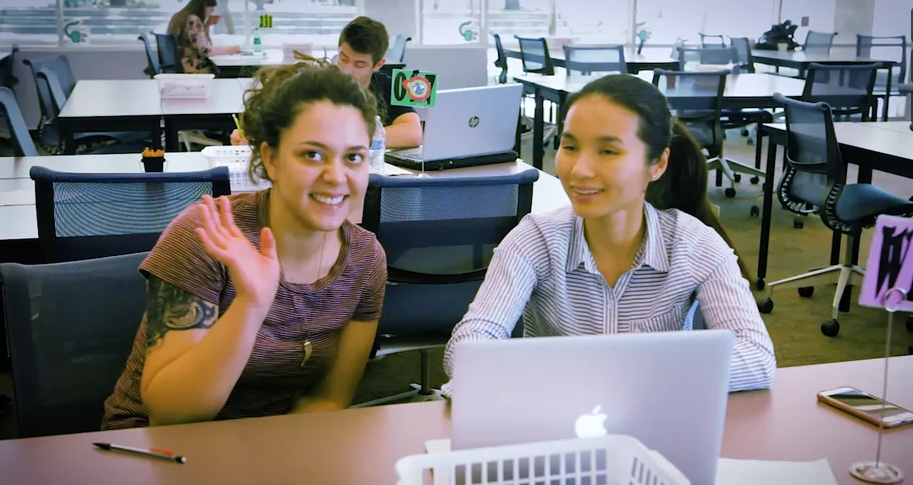 Two female students study at a computer. One of them is waving at the camera.