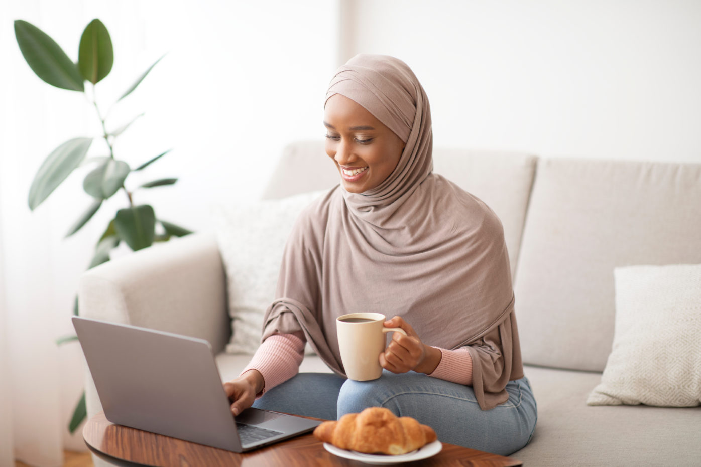 A female student wearing a hijab sits on a white couch while looking at a laptop on the coffee table in front of her. She is holding a mug in her left hand and is using the laptop with her right hand. There is a croissant on the coffee table and a green plant behind the couch.