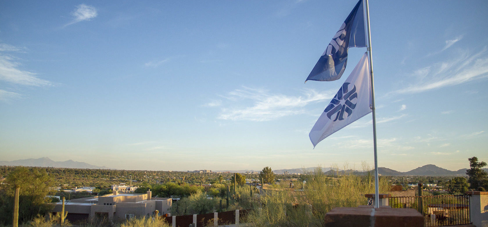 A blue and a white flag, both featuring the Pima Community College logo fly above Tucson