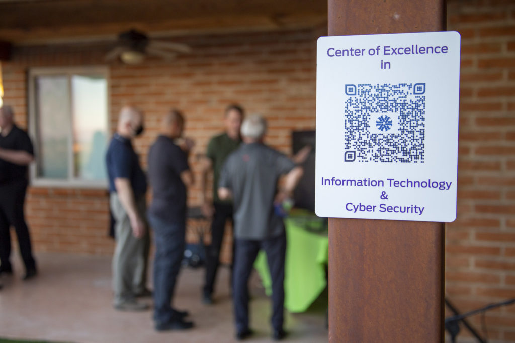 A sign with the QR code for the Center of Excellence in Information Technology and Cyber Security is in focus as a group behind it discusses the center.